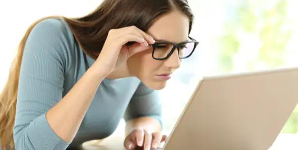Woman squinting and lifting her glasses to see a laptop screen, indicating astigmatism.