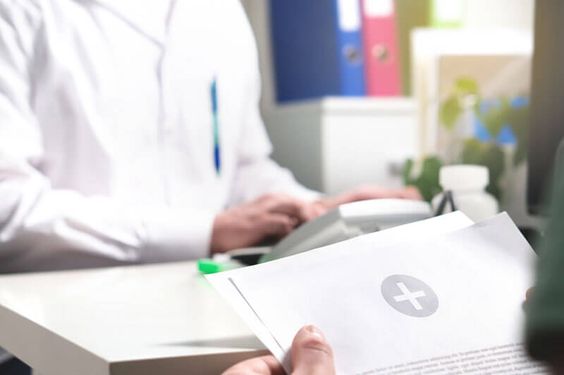 Close up of some medical paperwork held by a patient sitting in front of a desk, behind which sits a man in a white lab-coat.