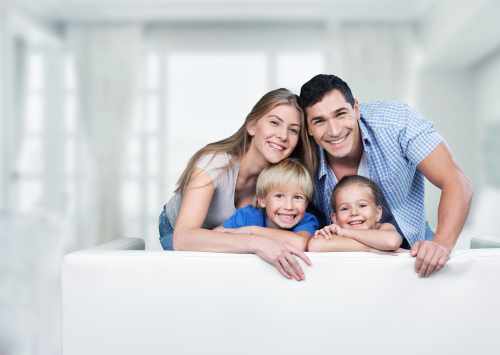 Family of four posing looking over the back of a white couch. Mom, dad and two young sons are smiling.