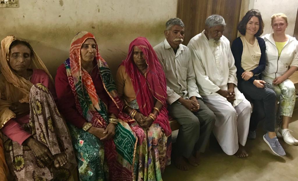 A group of elderly individuals in India, some with their eyes closed due to cataract-related vision impairment, seated alongside healthcare volunteers from Moorfields Eye Hospital Dubai
