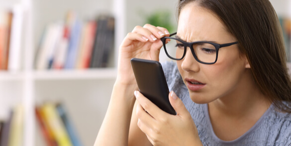 Woman with long dark hair and glasses squinting and holding a smartphone very close to her face, suggesting difficulty focusing on near objects, a sign of hypermetropia (farsightedness).