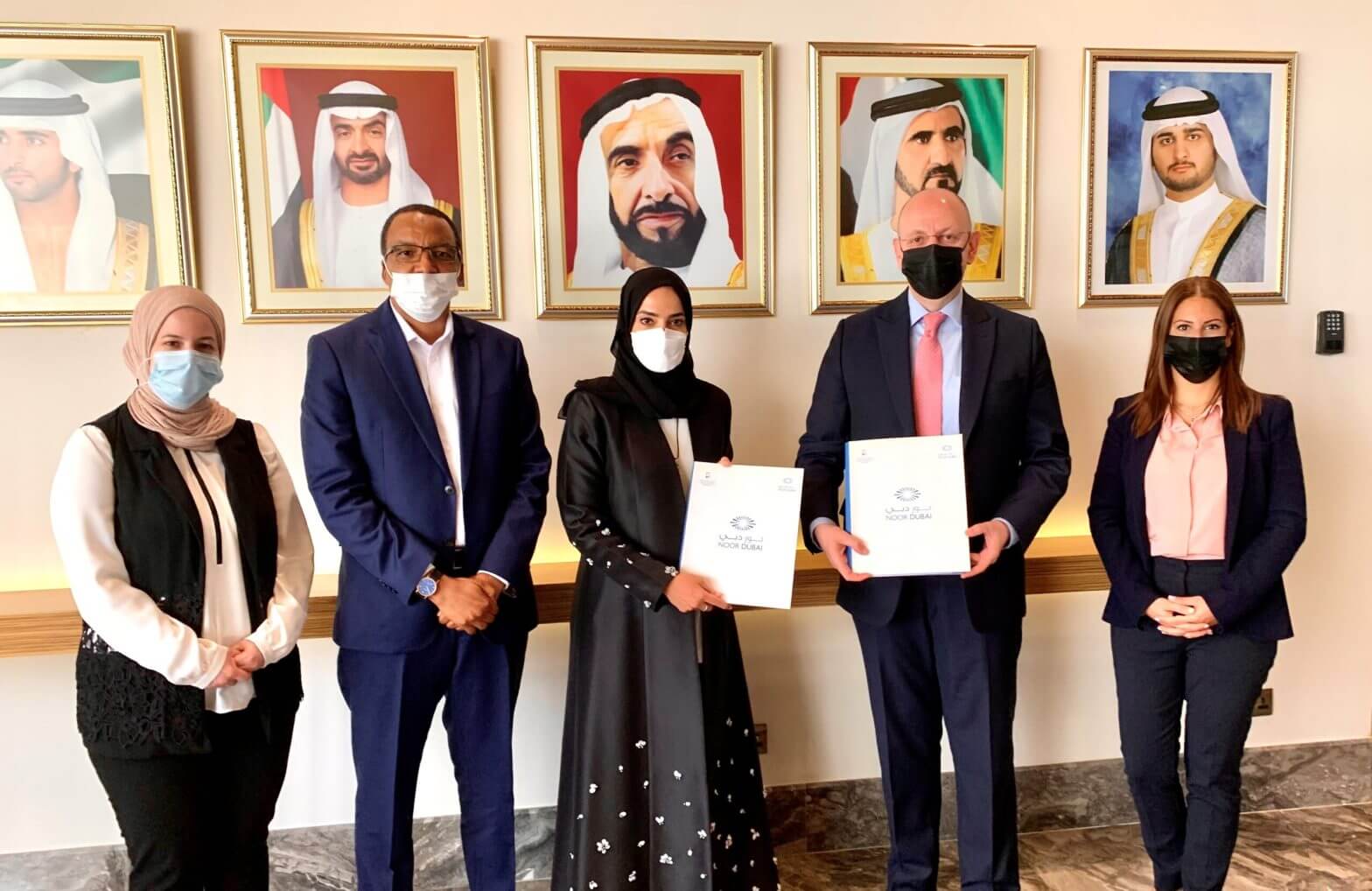 Image of Five people standing side by side, wearing medical masks, in front of a row of portraits on the wall. In the centre, a woman and a man standing together each hold a certificate.