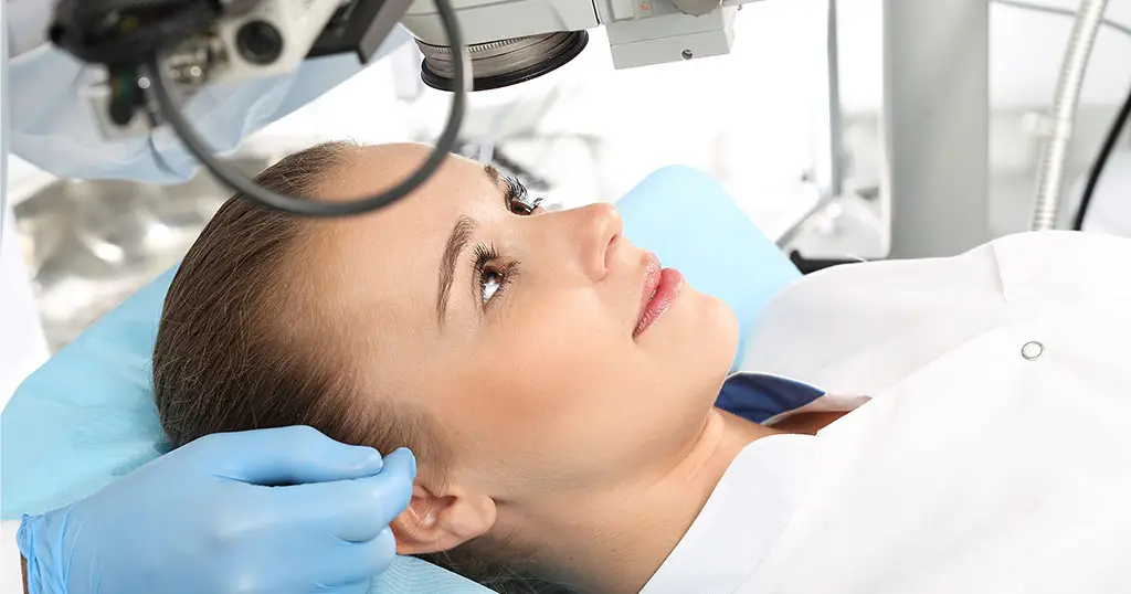 Woman lying under an ophthalmic microscope, preparing for an eye examination or laser eye surgery, with a gloved hand supporting her head.