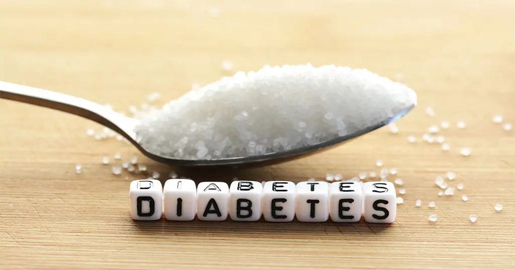 A spoon filled with white sugar granules placed on a wooden surface, with small cube beads in front spelling out the word “DIABETES.”
