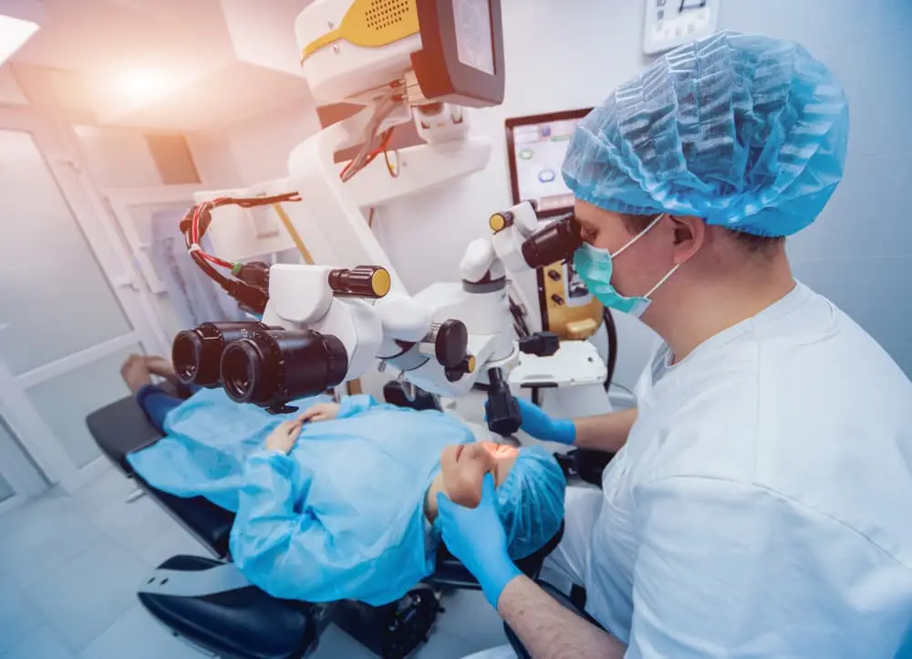 Surgeon in surgical scrubs and mask performing an eye procedure on a patient lying under an operating microscope in a modern clinic.