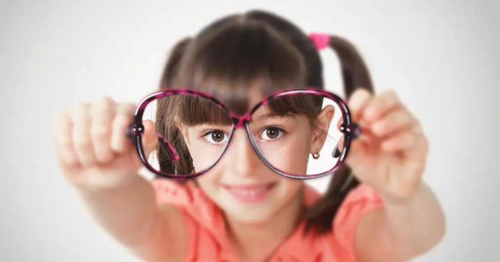 Young girl holding oversized glasses towards the camera, smiling and looking through the lenses indicatiing how glassess help her see better